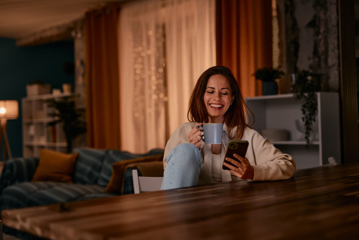 Una mujer toma café caliente relajada en su casa mientras revisa las notificaciones del móvil.