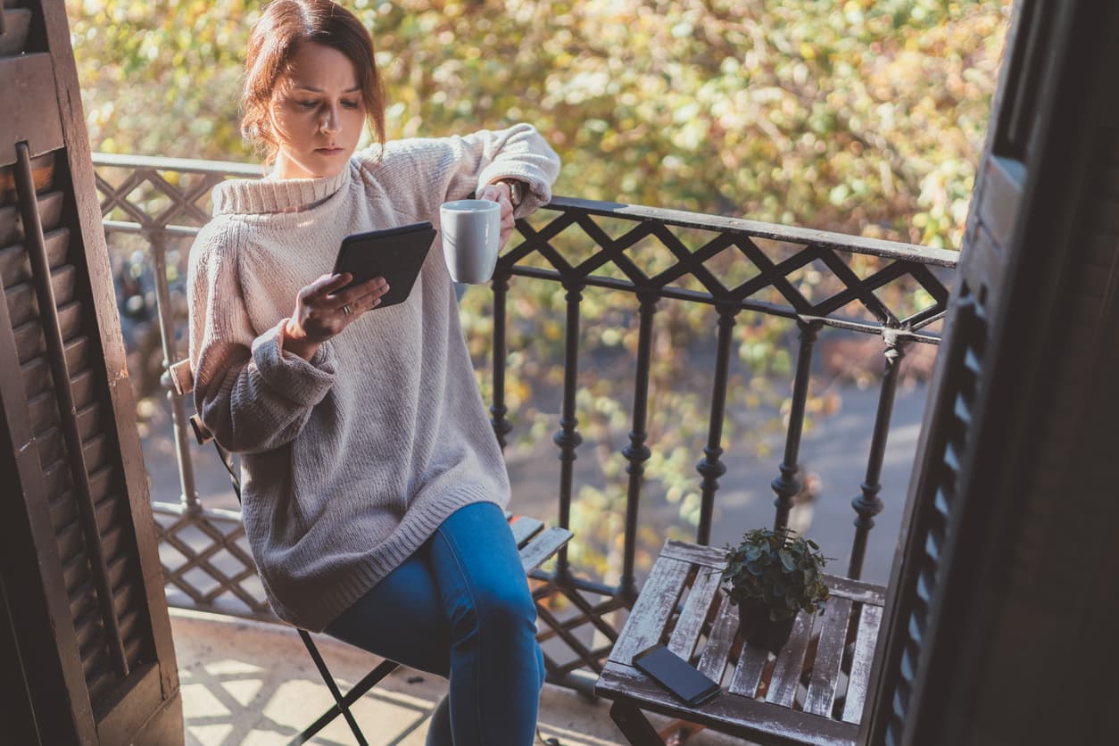 Una mujer lee un eBook en el balcón de su casa mientra toma café.