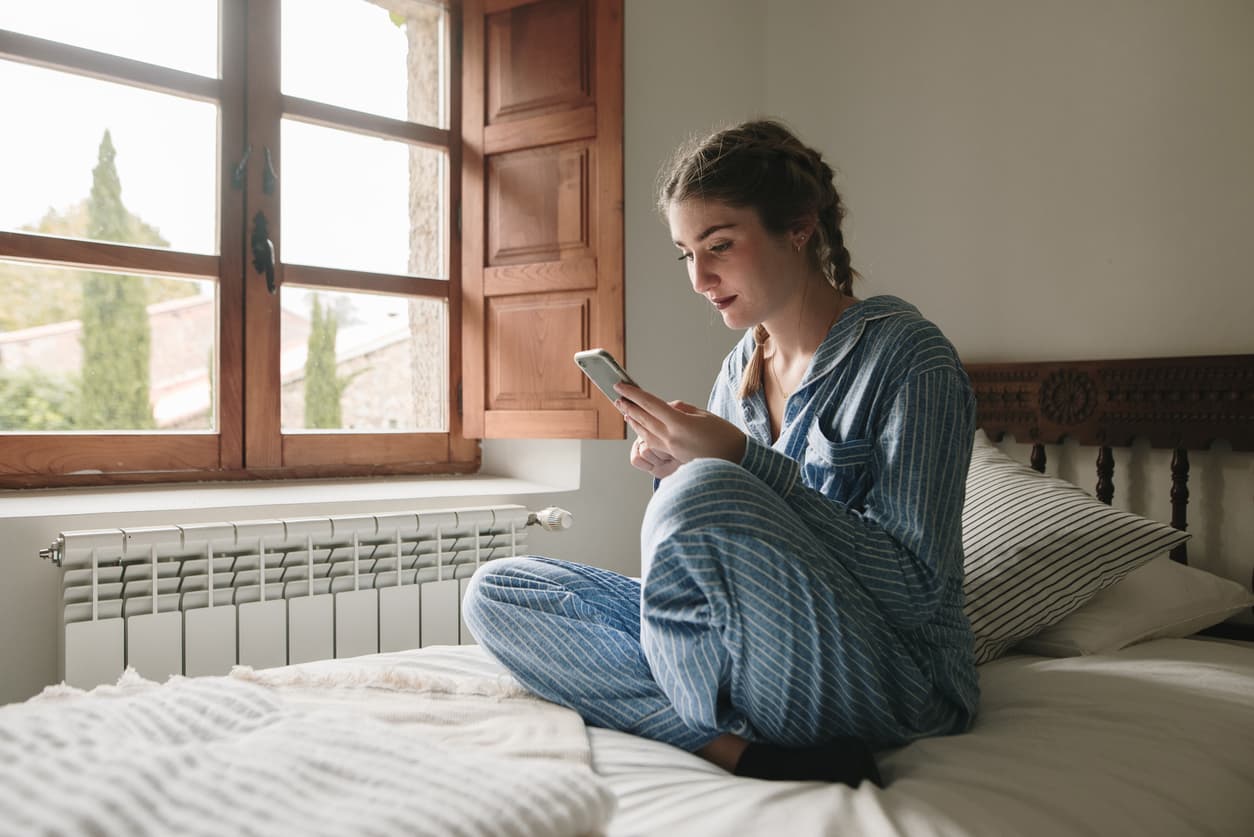 Una mujer mira el móvil mientras se protege del frio con un radiador de cerámica pequeño.